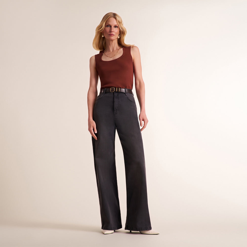 A woman stands against a plain warm beige studio background wearing a sleeveless rust-brown top, dark charcoal wide-leg pants with a thin brown belt, and pointed flats.