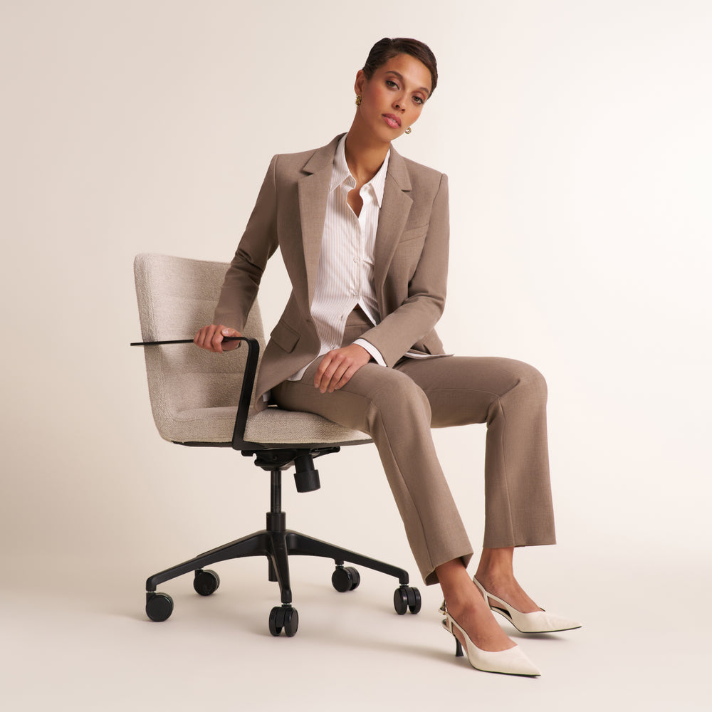 A woman sits on a light beige office chair wearing a tailored taupe suit with a white shirt and white pointed heels, photographed in a clean studio setting.