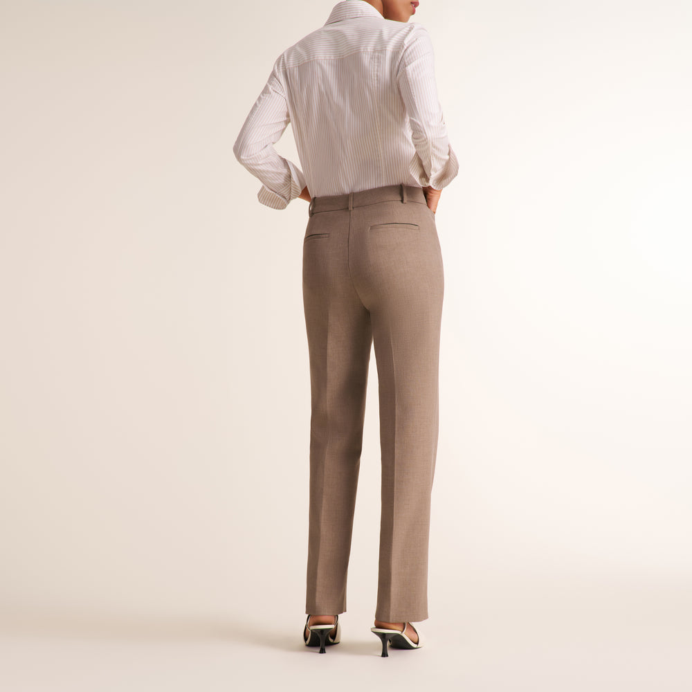 A woman shown from the back wearing straight-leg taupe trousers, a white button-down shirt, and black high heels against a plain cream studio background.