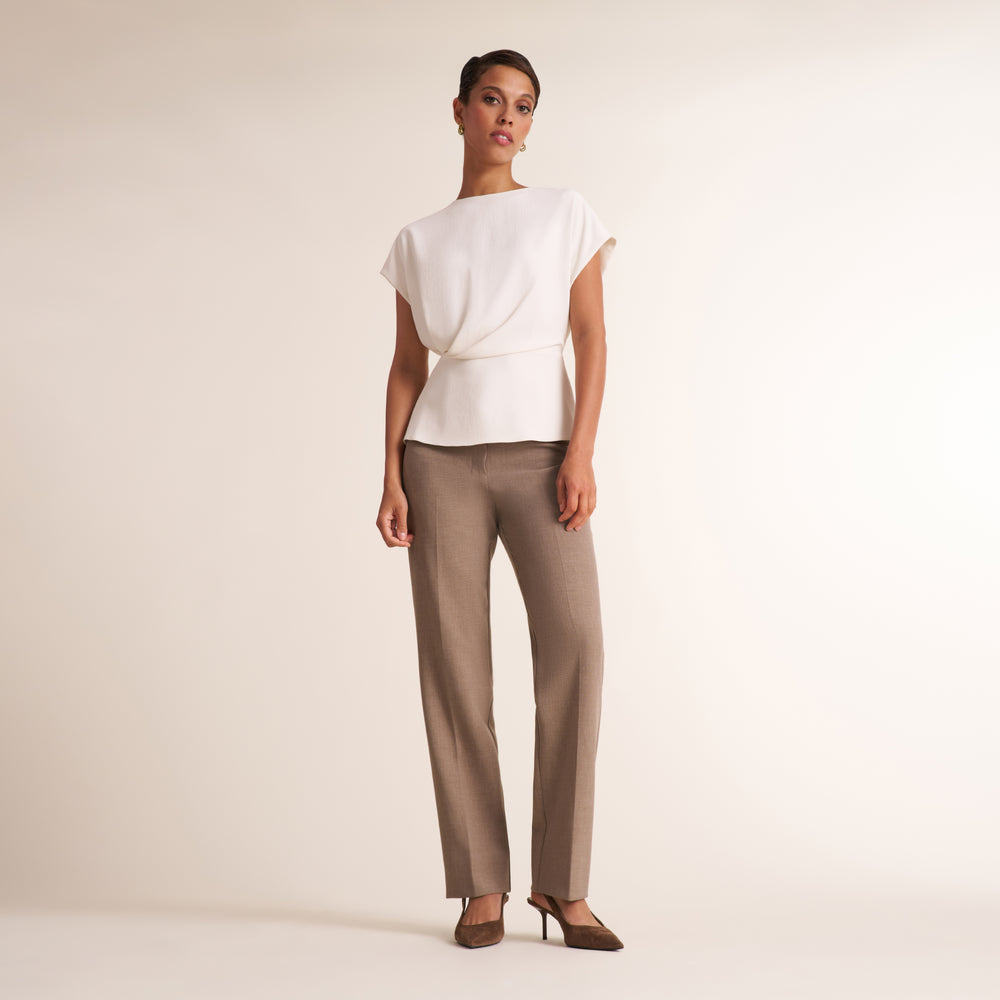 A model stands against a plain light beige studio background wearing a white draped short-sleeve top and taupe straight-leg trousers with brown heels.