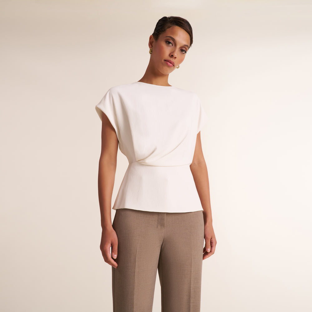 A woman modeling a white short-sleeve peplum top with a draped wrap-style bodice, paired with taupe trousers, against a plain light beige studio background.