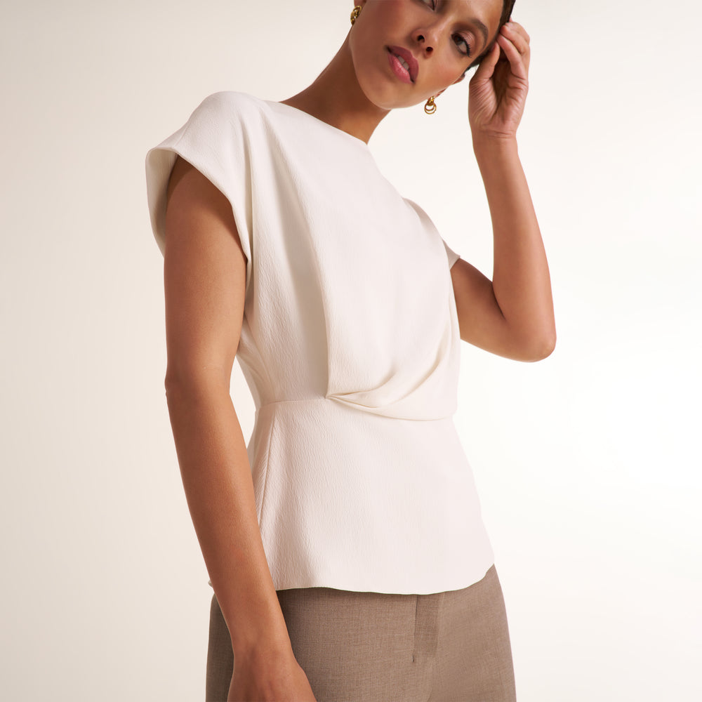 A woman wearing a cream-colored short-sleeve top with a draped, asymmetrical front detail and a peplum-like waist, paired with taupe trousers, against a plain light background.