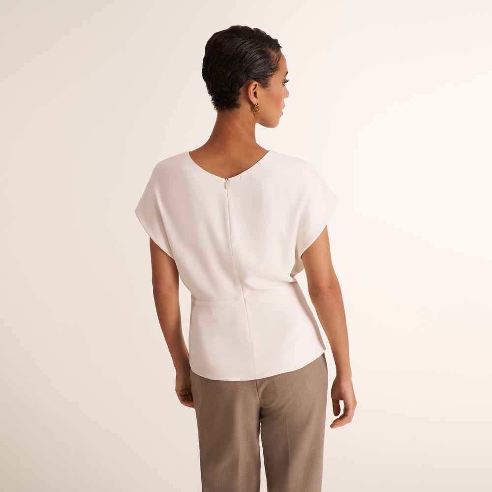 A woman shown from the back wearing a cream-colored short-sleeve top with a V-neckline and a zippered back, paired with beige trousers, against a plain cream studio background.