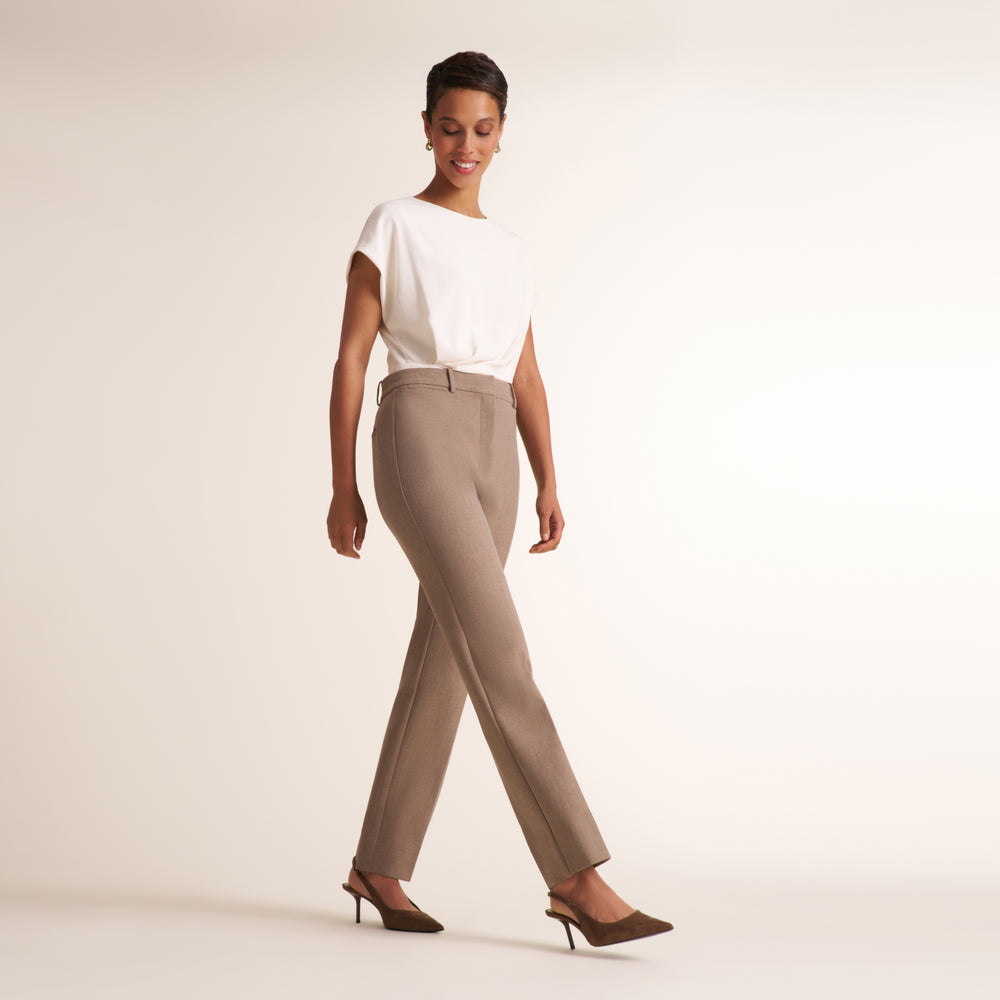 A woman modeling high-waisted, straight-leg trousers in a light taupe/beige color, paired with a white short-sleeve top and dark brown slingback heels, against a solid off-white studio background.