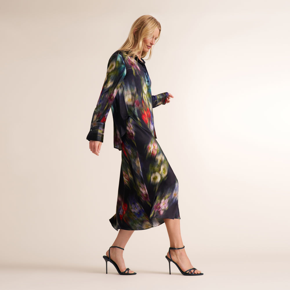A woman models a long-sleeved midi dress with a dark navy base and a vibrant, painterly multicolor floral print. She is wearing black strappy high-heeled sandals against a plain, warm off-white studio background.