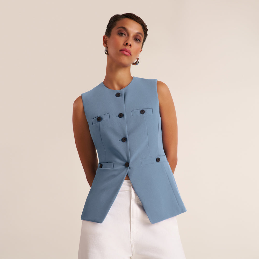 A woman wearing a sleeveless light blue vest with black buttons over white pants, photographed against a plain beige studio background.