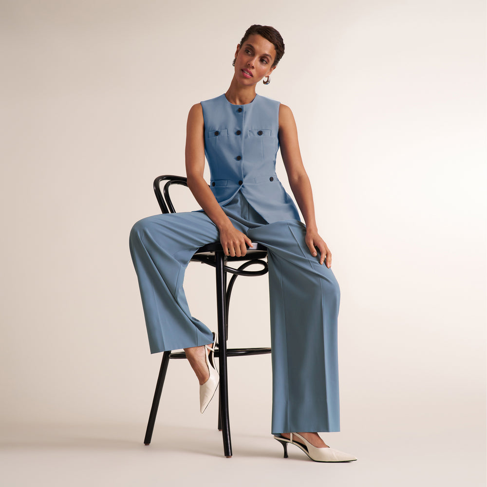 A model wearing a light blue sleeveless button-front top and matching wide-leg pants, seated on a black metal stool against a plain light beige studio background.