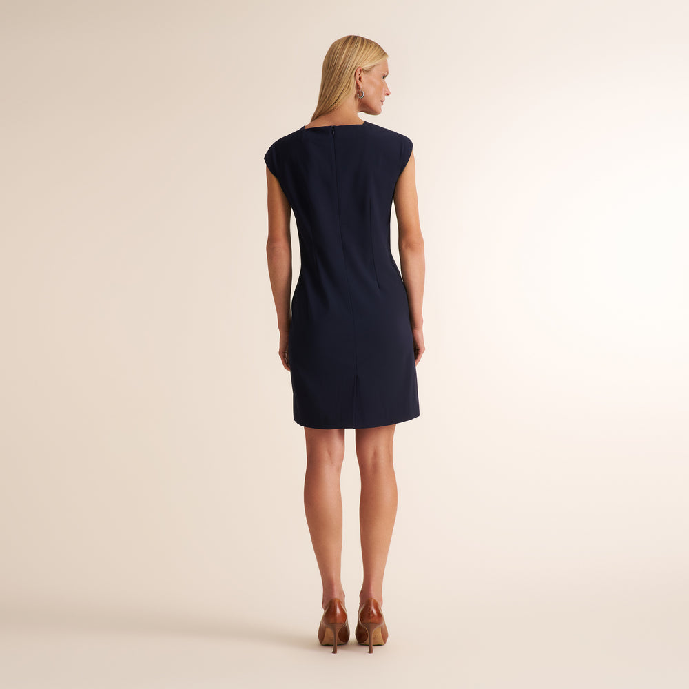 A woman viewed from the back wearing a sleeveless navy blue shift dress and brown high heels, standing against a plain light beige studio background.