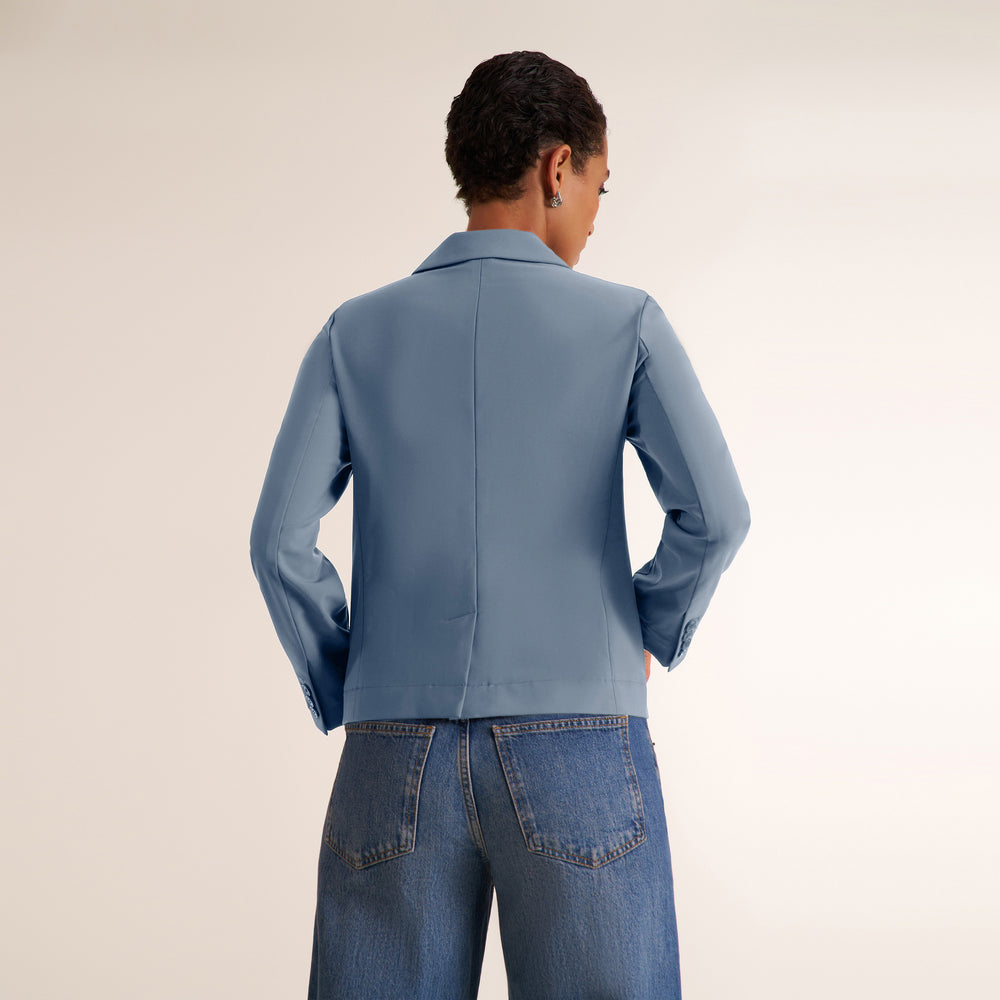 A model shown from the back wearing a dusty blue cropped blazer and blue jeans against a plain light beige studio background.