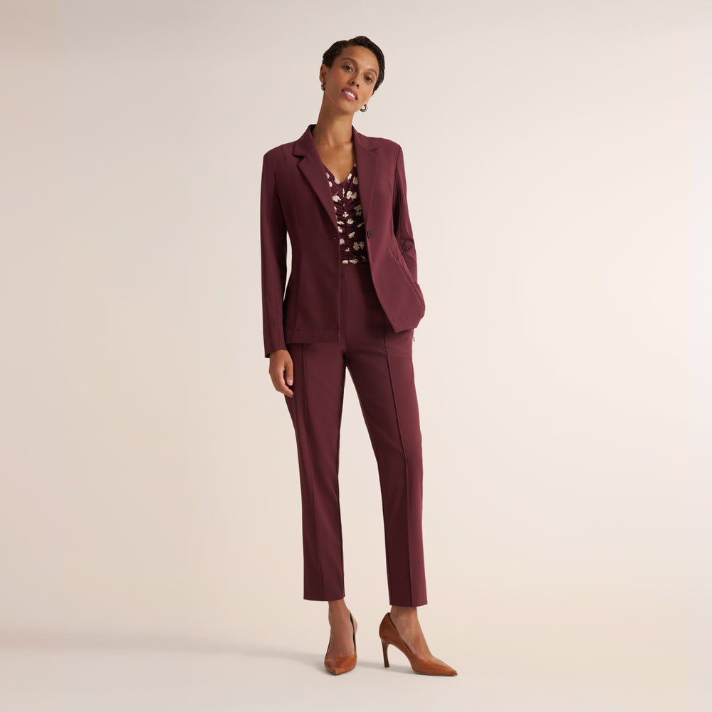 A woman modeling a matching burgundy two-piece suit with a patterned blouse and brown heels, standing against a plain beige studio background.