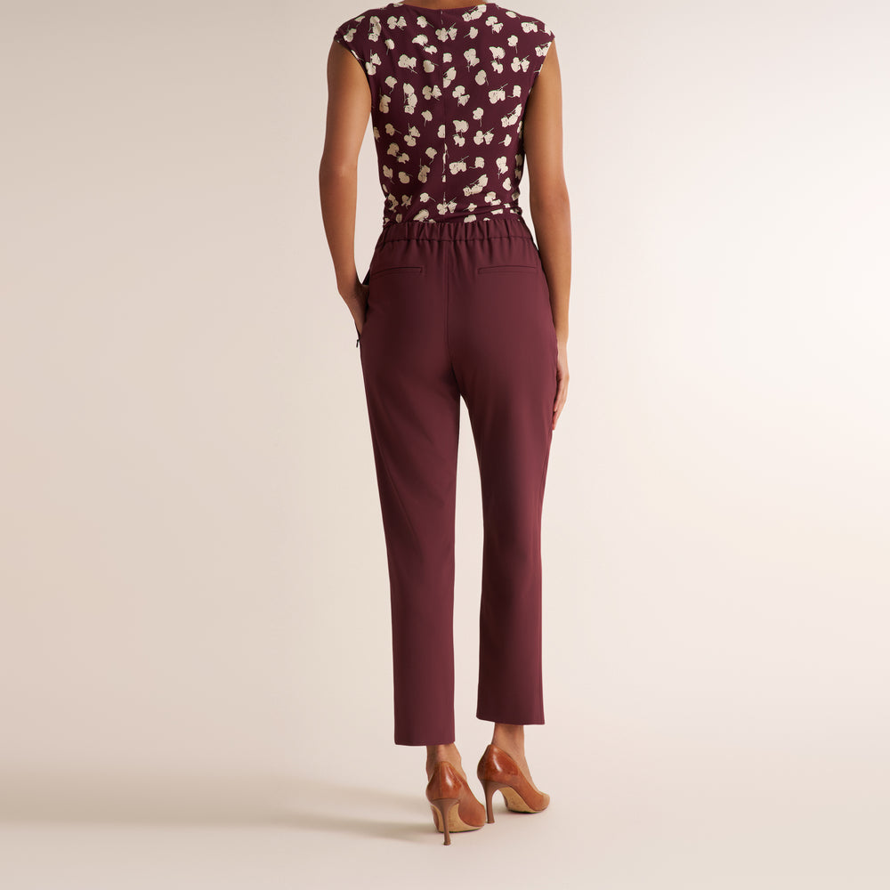 A model shown from the back wearing a sleeveless burgundy top with a white floral pattern and matching burgundy cropped trousers, paired with tan high heels, against a solid light beige background.