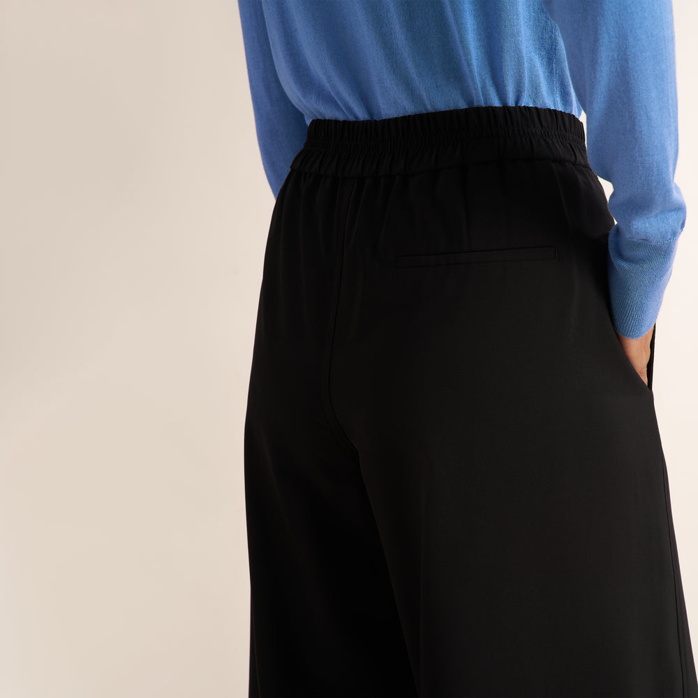 A model shown from the back wearing black wide-leg cropped pants, a blue long-sleeve top, and black high-heeled sandals against a plain beige studio background.
