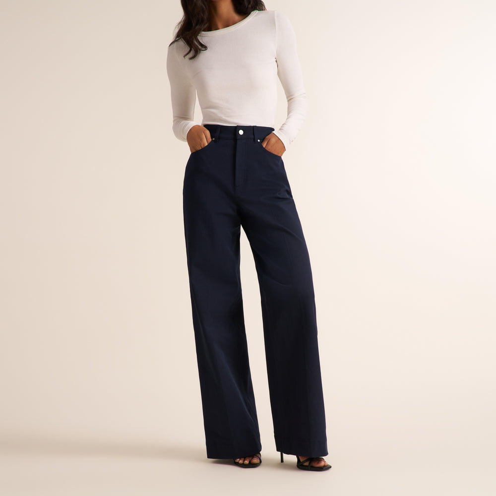 A model wearing navy blue wide-leg trousers and a white long-sleeve top, standing against a solid light beige background.
