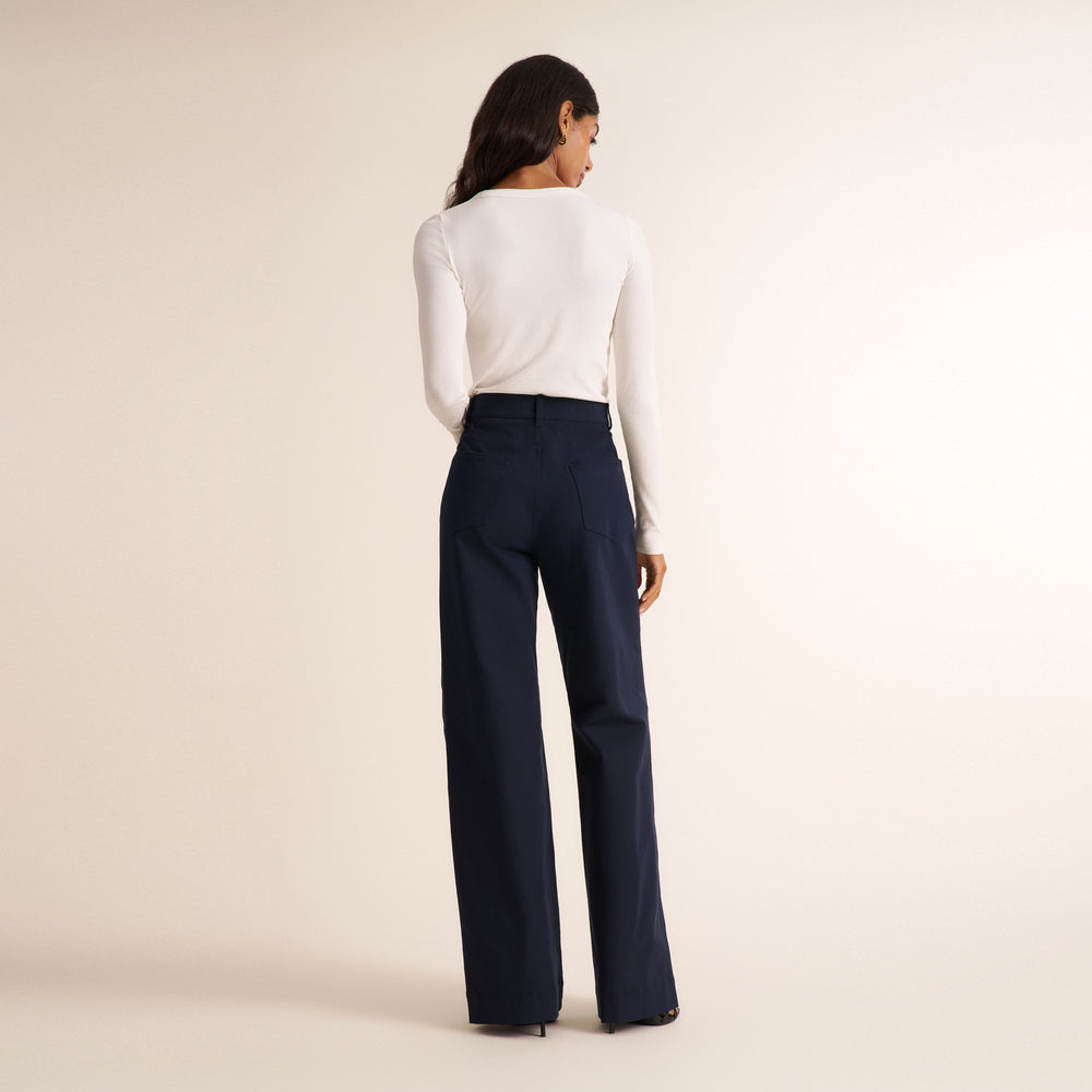 A woman is shown from behind wearing a fitted white long-sleeve top and high-waisted navy blue wide-leg trousers, standing against a plain beige studio background.