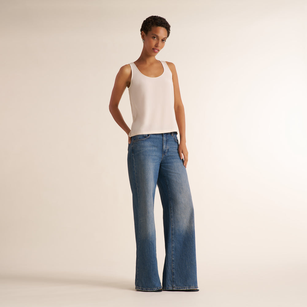 A woman modeling a sleeveless scoop-neck tank top in a light cream color, paired with blue jeans, against a plain beige studio background.