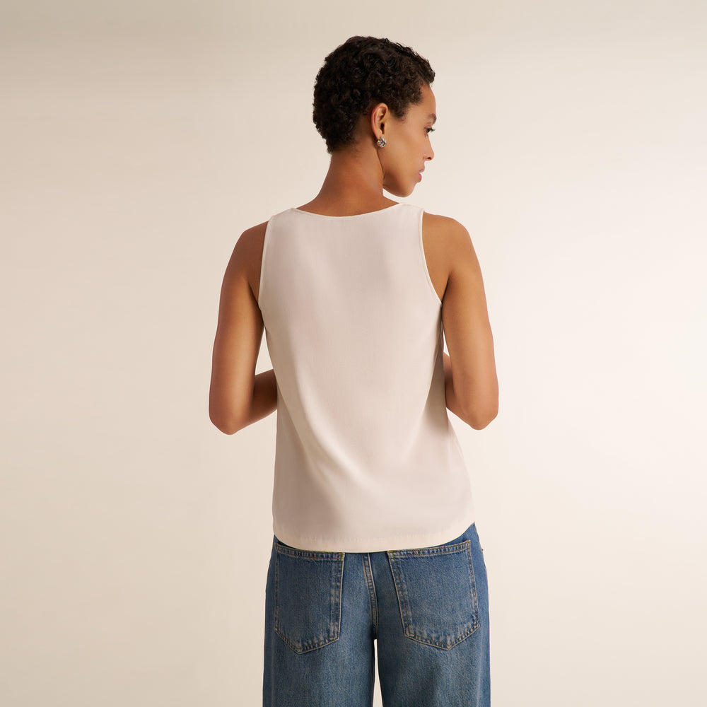 A woman shown from the back wearing a sleeveless cream-colored top and blue jeans, standing against a plain light beige studio background.