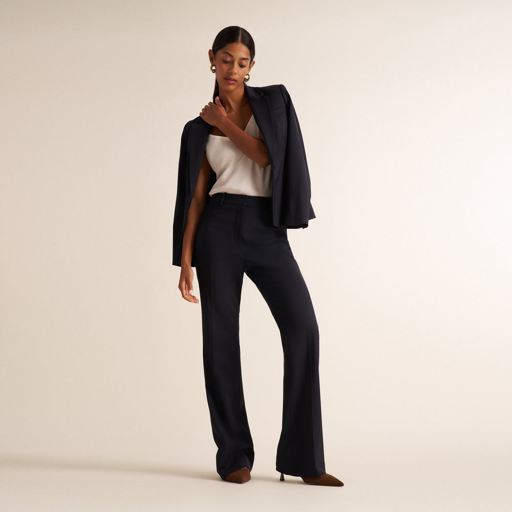 Woman wearing a black blazer, wide leg black suit pants, and a v-neck white tank top standing on a beige background