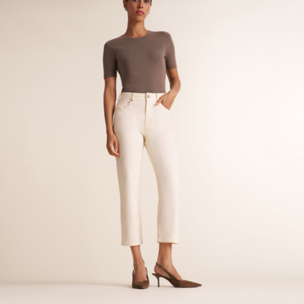 A model wearing a fitted taupe short-sleeve top, off-white cropped trousers, and brown pointed-toe heels, standing against a plain warm beige studio background.