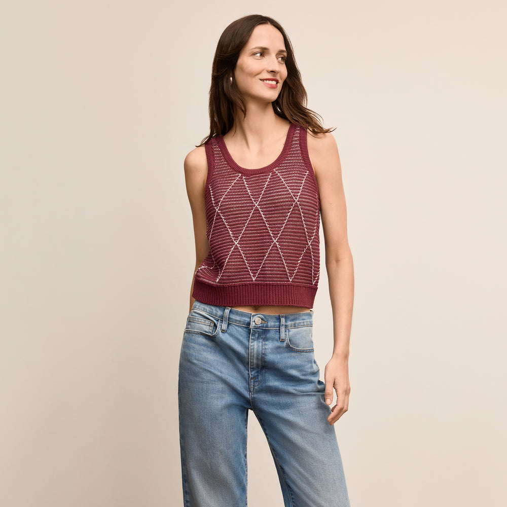 Woman wearing a maroon sleeveless knitted top and blue jeans on a beige background