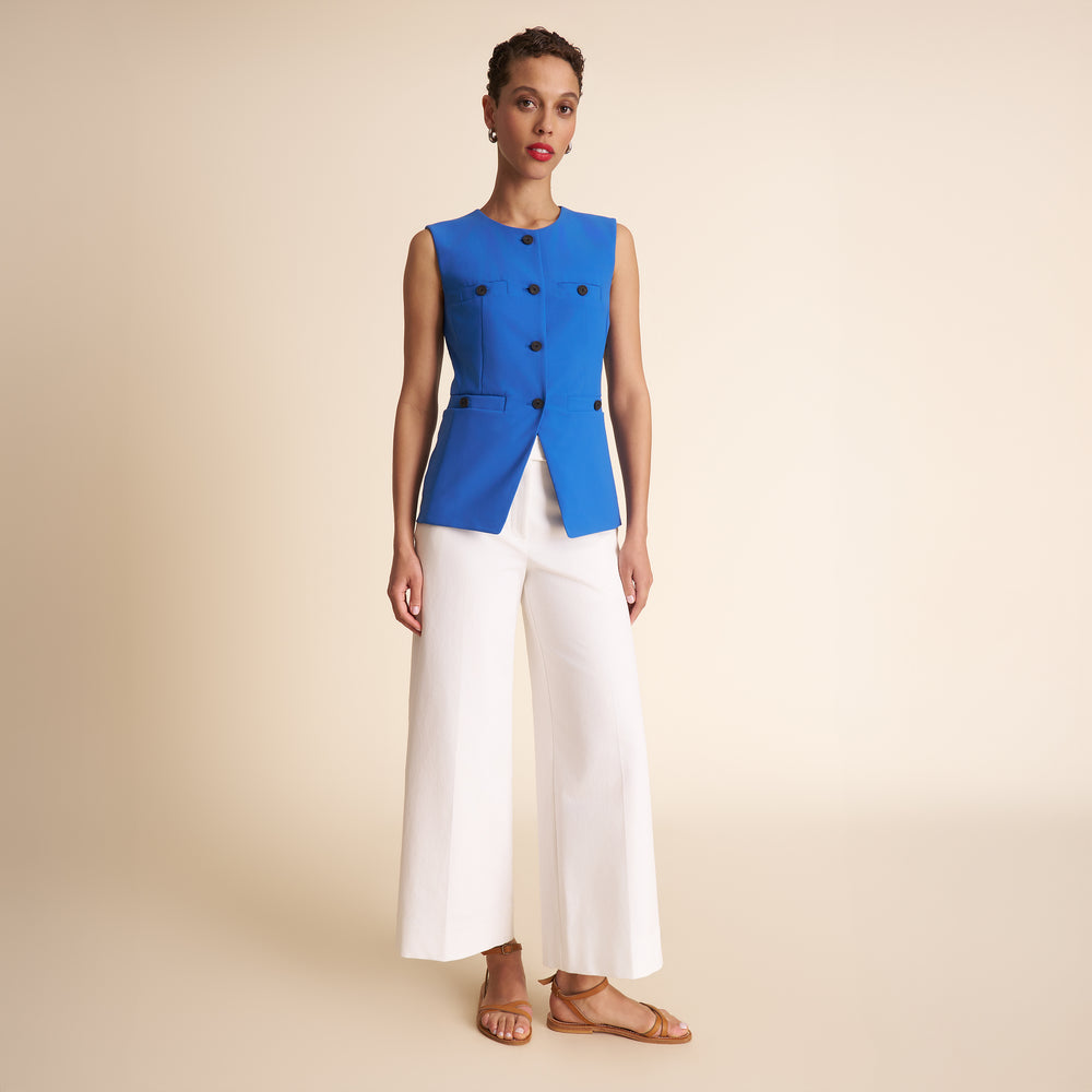 Woman wearing a blue sleeveless top with black buttons and white pants on a beige background