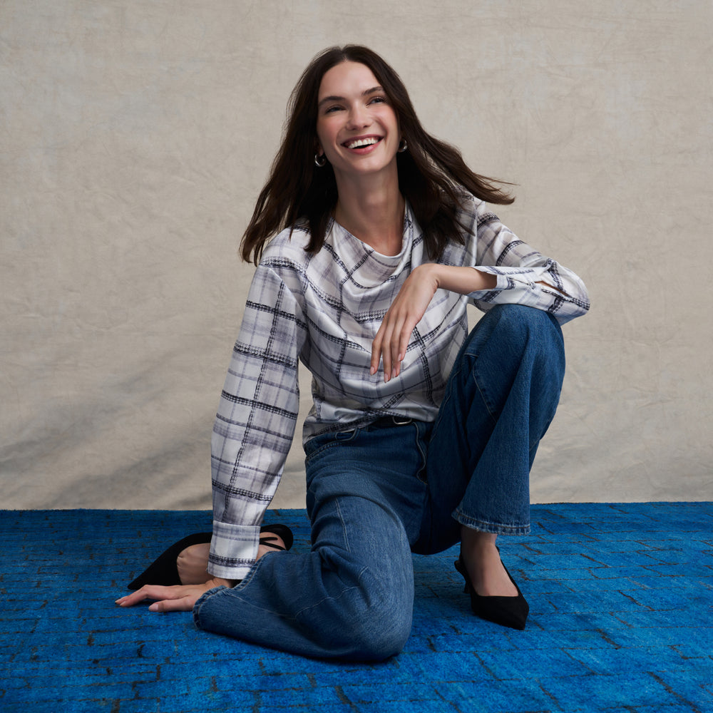 Woman wearing a white and gray plaid silk long sleeve boat neck top and wide leg jeans on a blue carpet floor and against a beige background