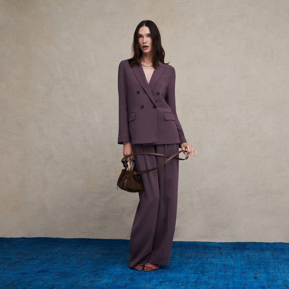 Woman wearing a purple hazy mauve double breasted blazer and matching wide leg pants on a blue carpet floor and against a beige background