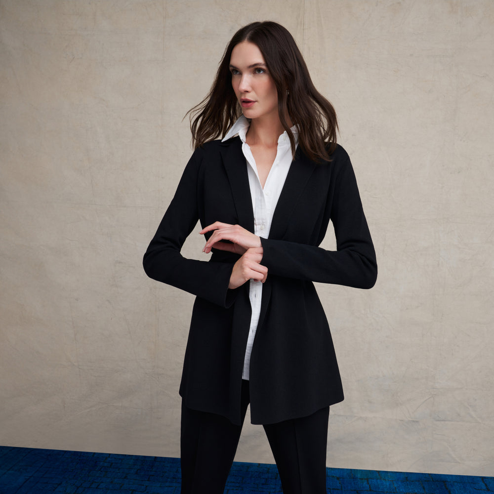 Woman wearing a black wrap jardigan, white button down shirt, and black skinny pants on a blue carpet floor and against a beige background