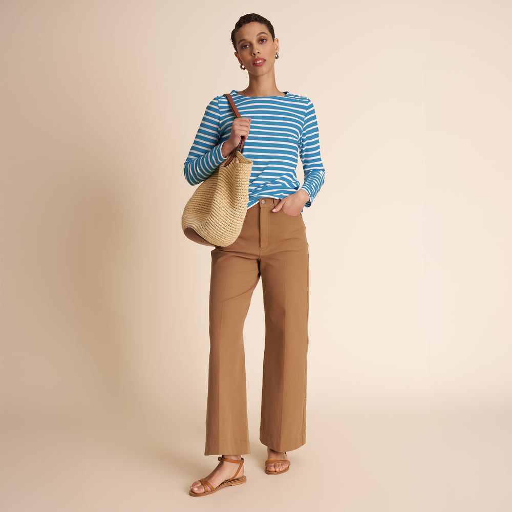 Woman wearing a blue striped shirt and tan pants, holding a straw bag on a beige background