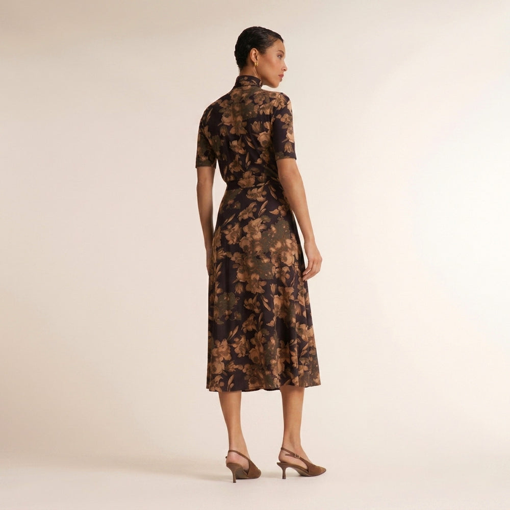 A woman stands with her back to the camera wearing a short-sleeved midi dress with a dark brown and black floral pattern, paired with brown heeled sandals, against a plain beige studio background.