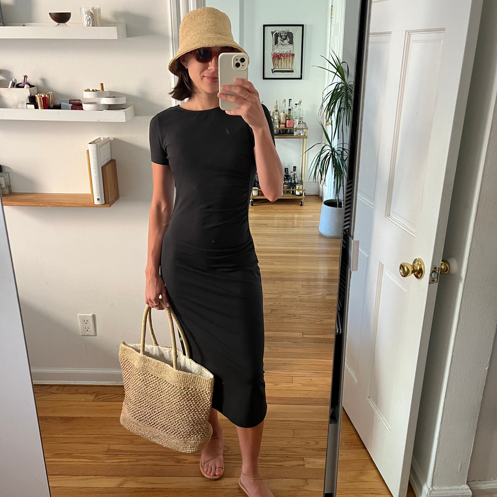 Person taking a mirror selfie wearing a black dress, beige bucket hat, and holding a woven bag.