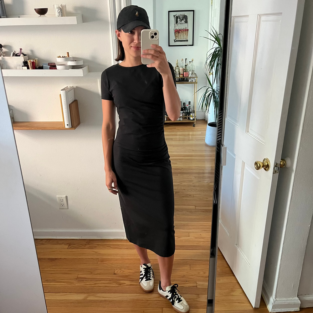 Person wearing a black dress and cap taking a mirror selfie in a room with wooden flooring and white walls