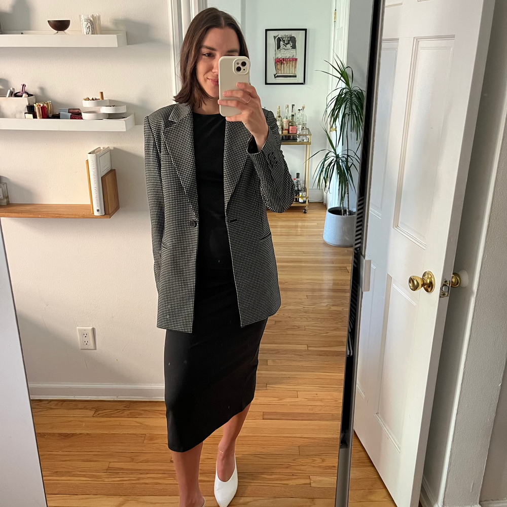 Person taking a mirror selfie wearing a gray blazer and black dress in a room with wooden flooring and white walls.