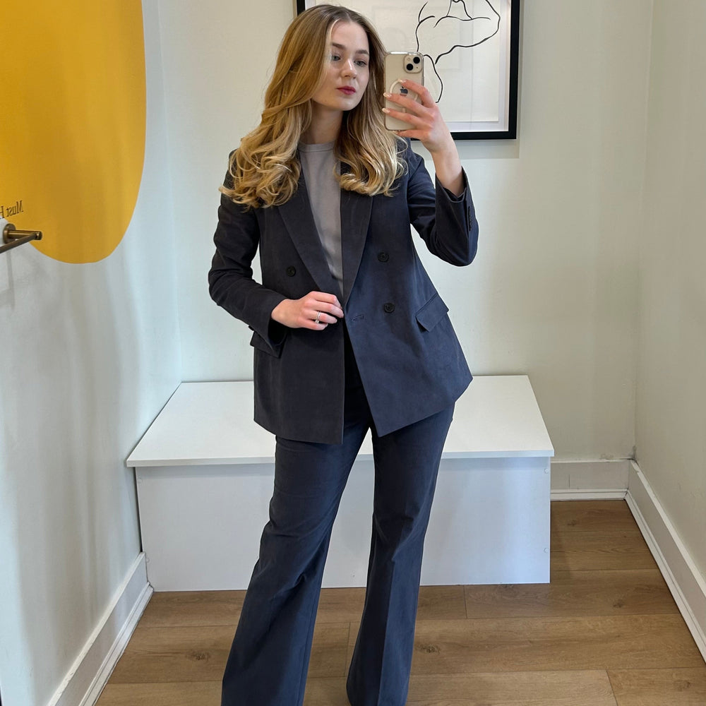 Woman taking a mirror selfie wearing a navy suit in a room with a white bench and framed picture on the wall.