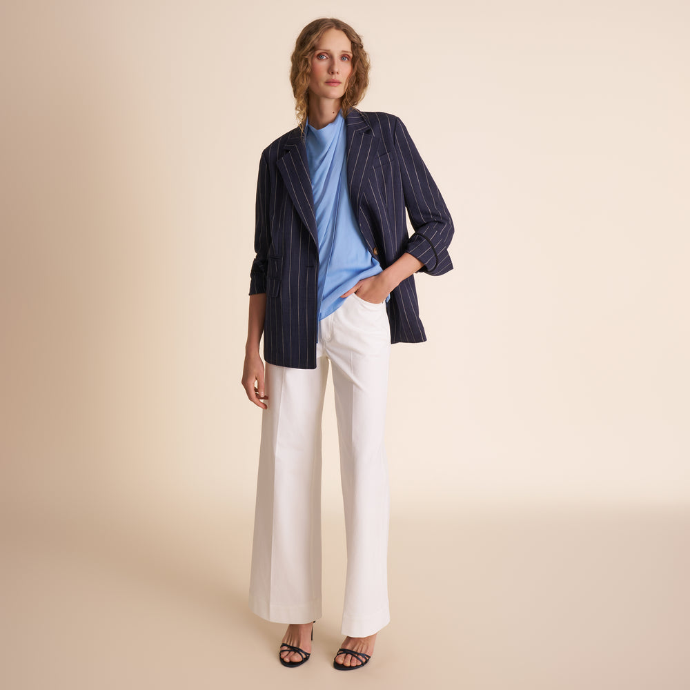 Woman wearing a navy striped blazer, blue shirt, and white pants on a beige background