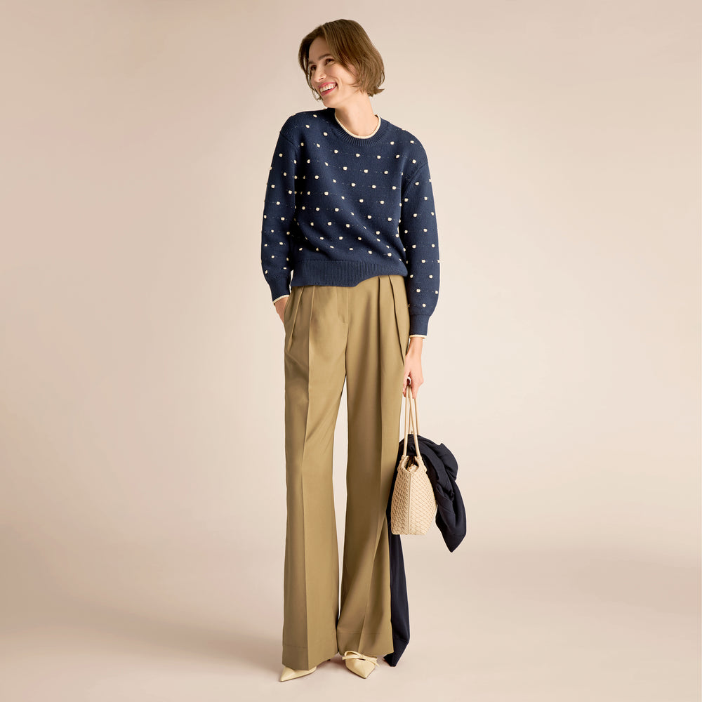 Woman wearing a navy sweater with white polka dots and beige pants, holding a beige bag on a beige background.