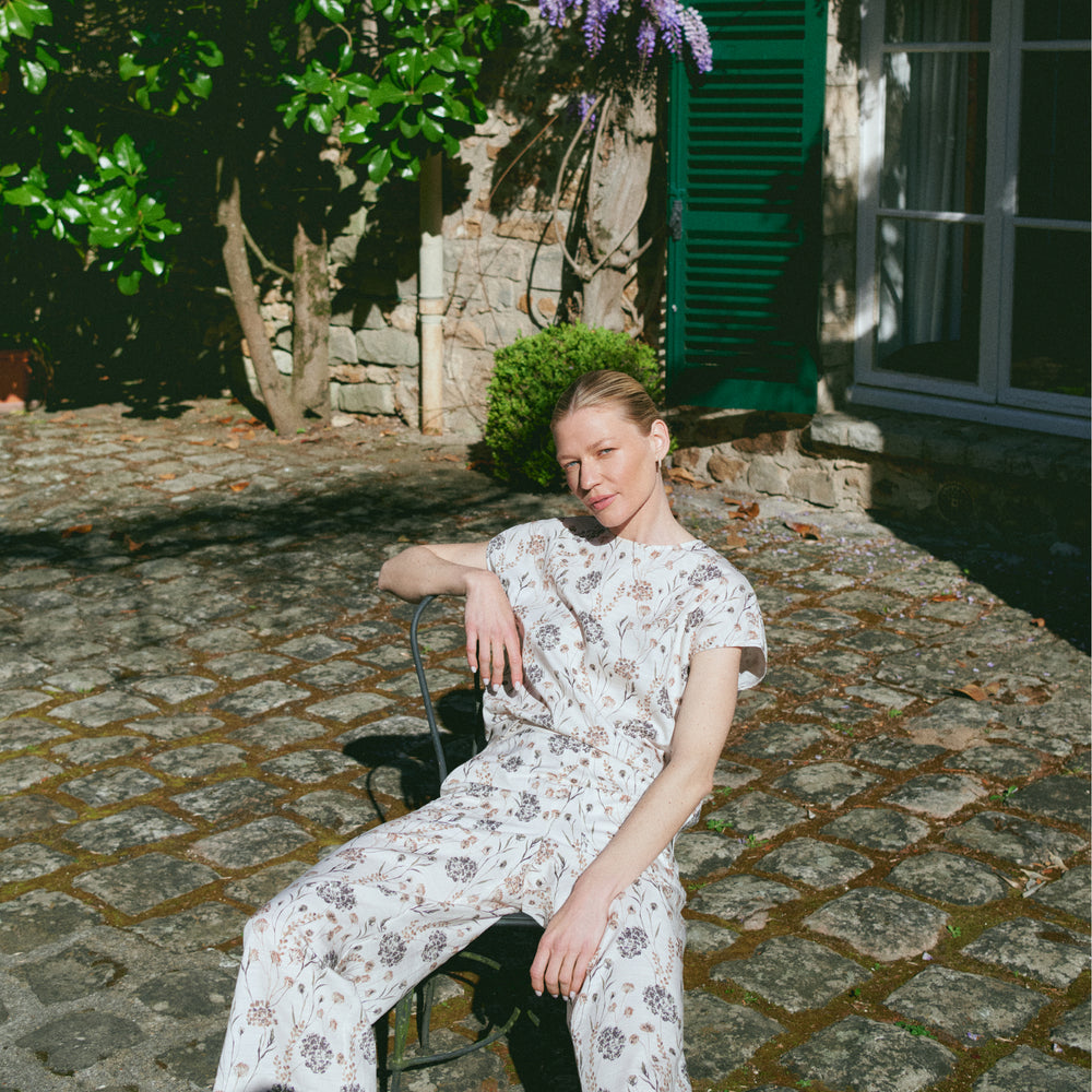 Woman in a floral dress sitting on a chair outdoors with greenery and a building in the background.