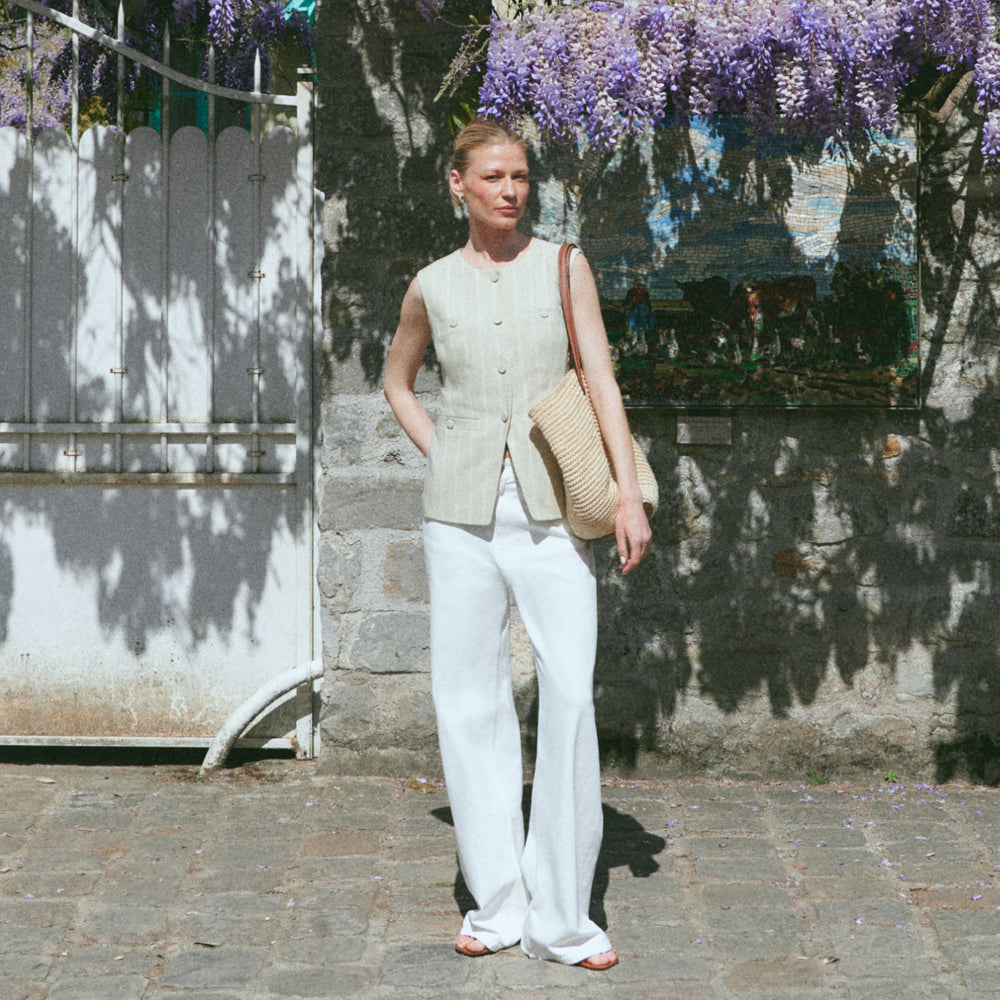 Woman in a light-colored sleeveless top and white pants standing against a stone wall with wisteria.