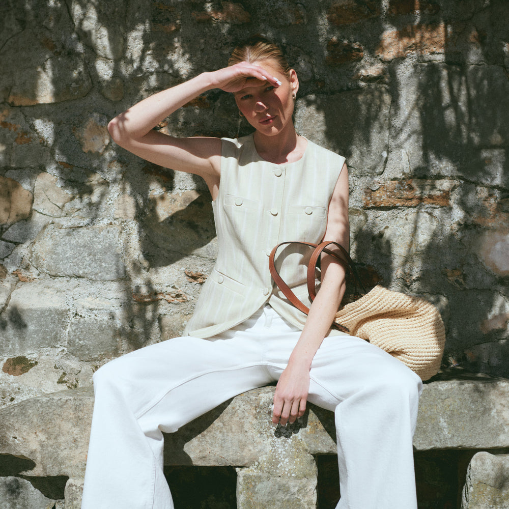 Woman sitting against a stone wall wearing a sleeveless top and white pants, holding a beige bag.