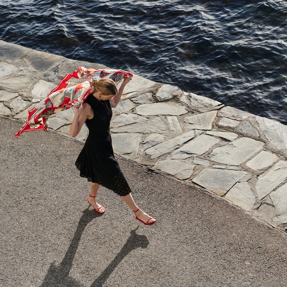 Woman in a black dress holding a red and white patterned scarf by a stone wall with water in the background
