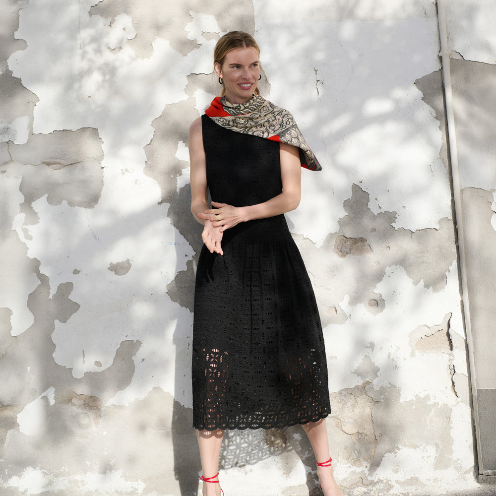 Woman wearing a black dress with a patterned scarf against a textured white wall.