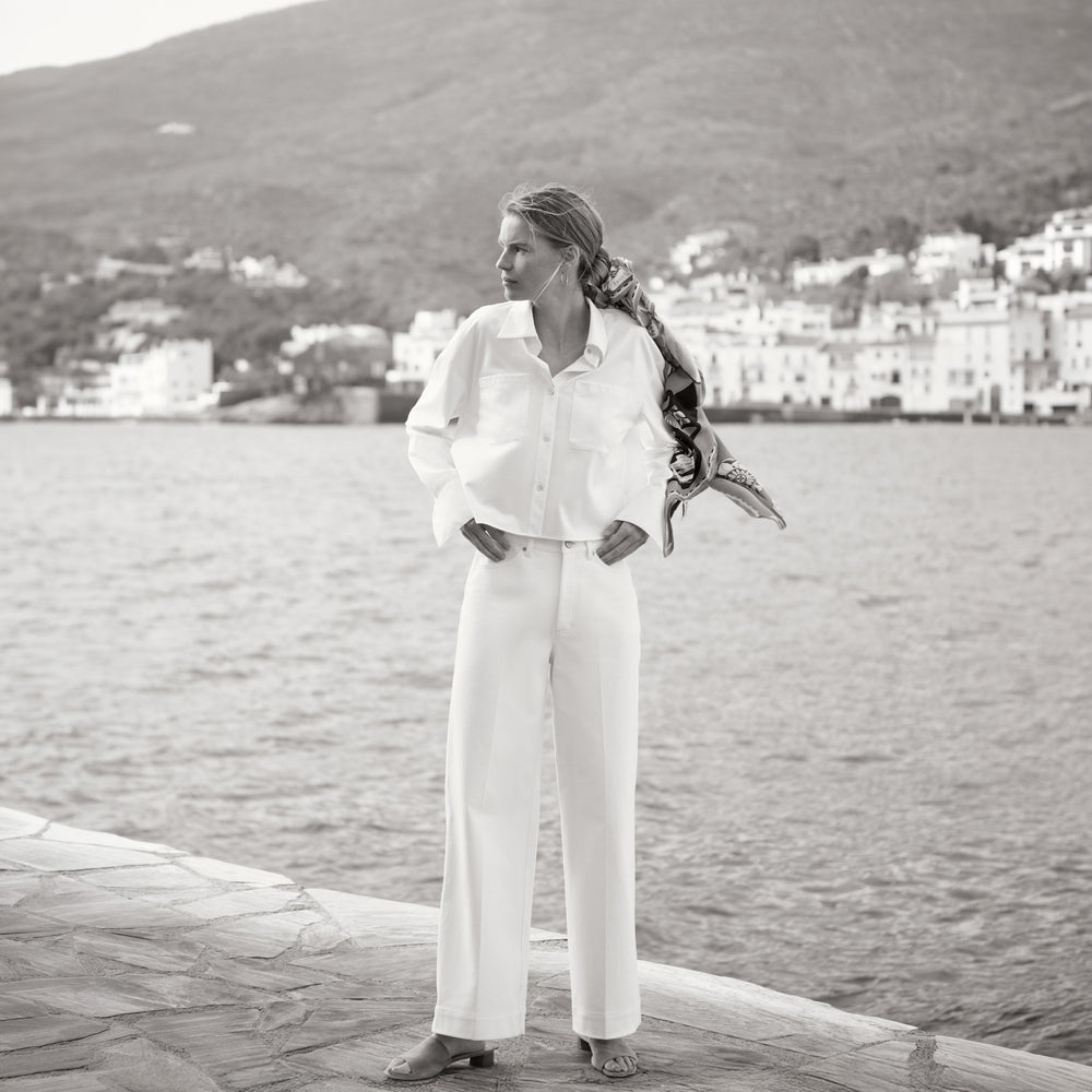 Woman in a white outfit standing by a waterfront with mountains in the background
