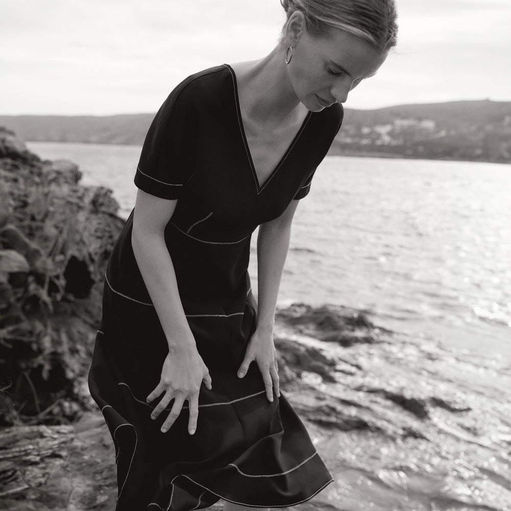 Woman in a black dress standing by a body of water with mountains in the background