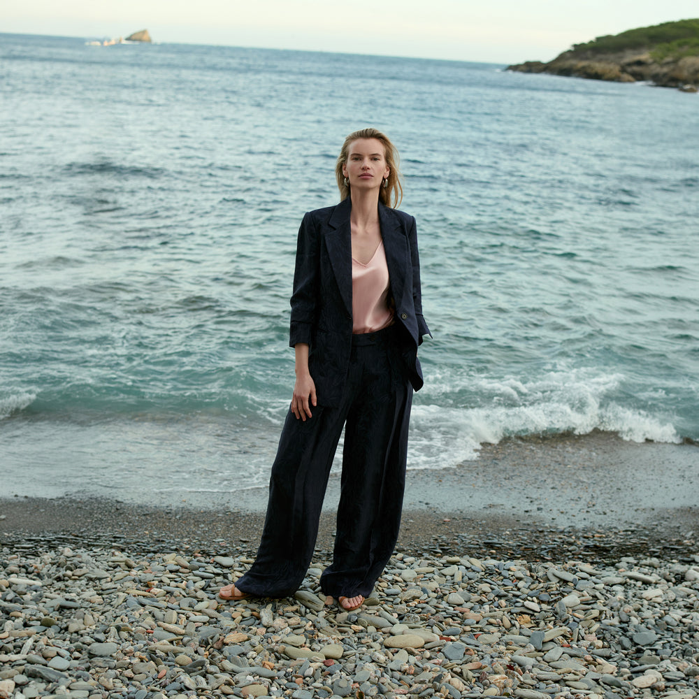 Woman in a black suit standing on a pebbly beach with ocean in the background