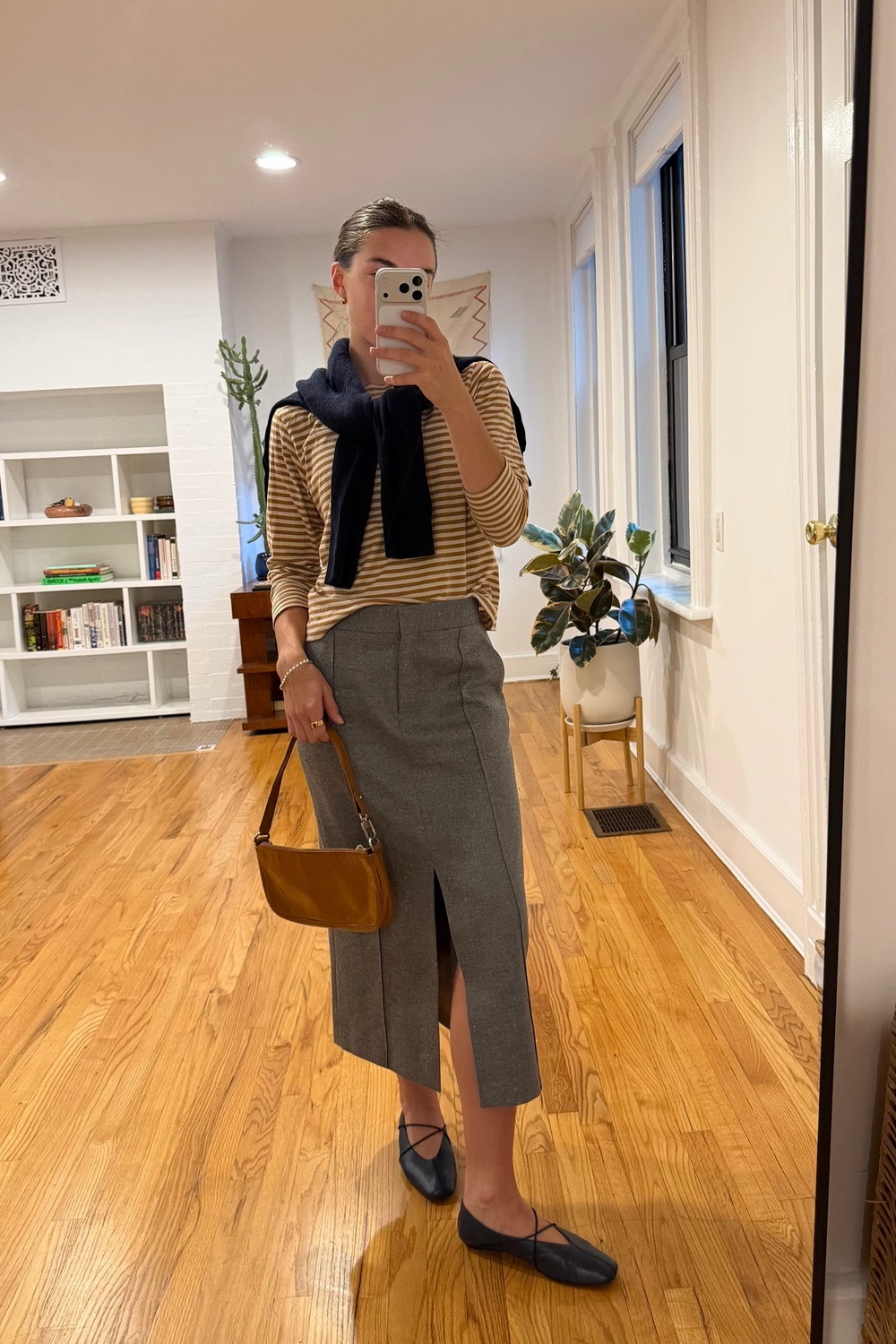 Person taking a mirror selfie wearing a striped sweater, gray skirt, and holding a brown bag in a room with wooden flooring.