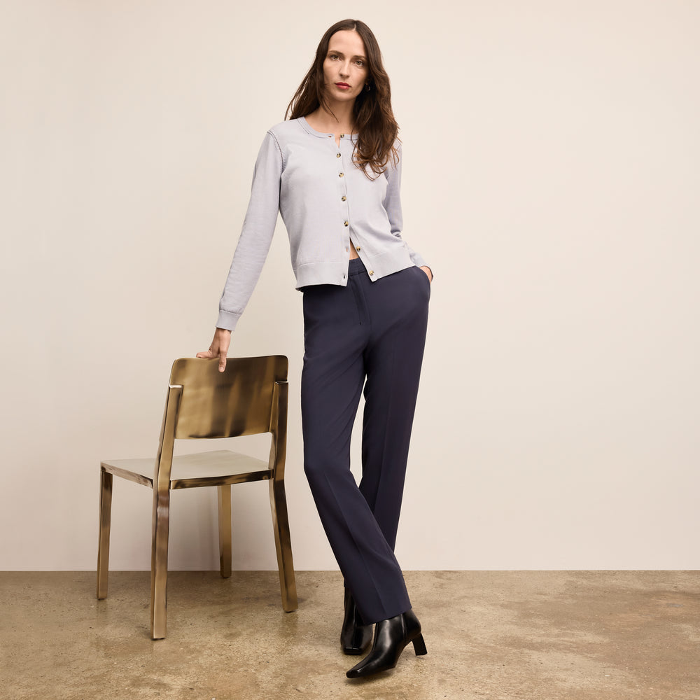 Woman in a light gray cardigan and navy pants standing next to a gold chair against a beige wall.