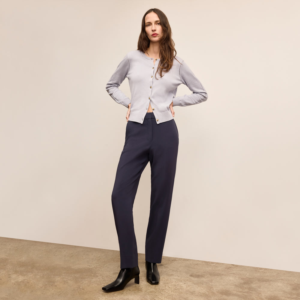 Woman wearing a light gray cardigan and dark blue pants against a beige background