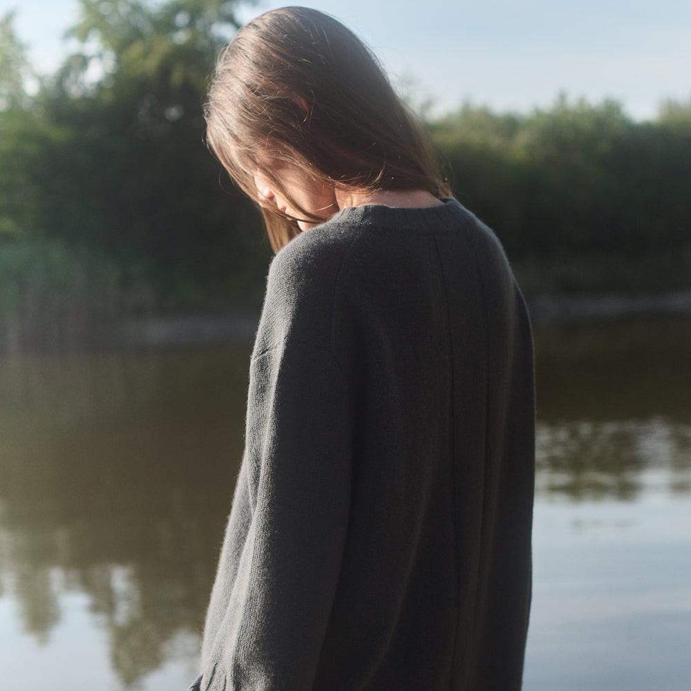 Person wearing a dark sweater standing by a body of water with trees in the background