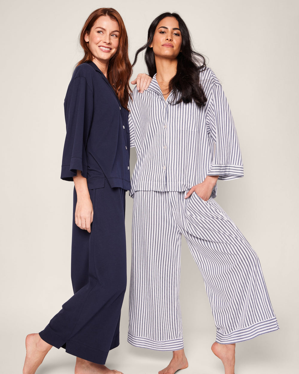 Two women wearing navy and white striped pajama sets on a plain background