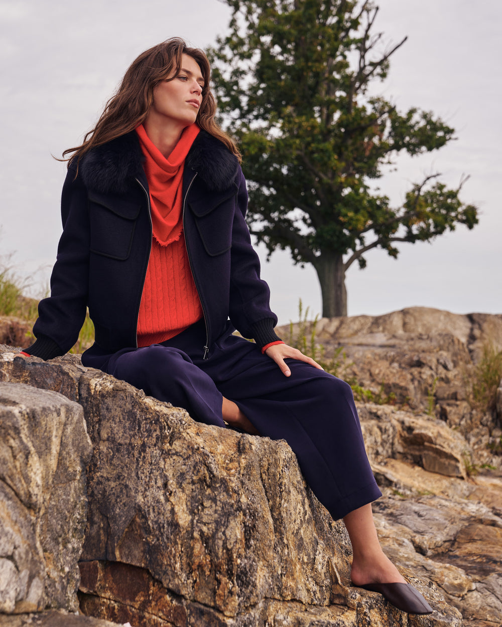A woman sits on a rocky outcrop outdoors, wearing a dark navy jacket with a dark fur collar, a bright orange turtleneck sweater, matching dark navy wide-leg pants, and dark brown pointed mule flats. A tree is visible in the background under an overcast sky.