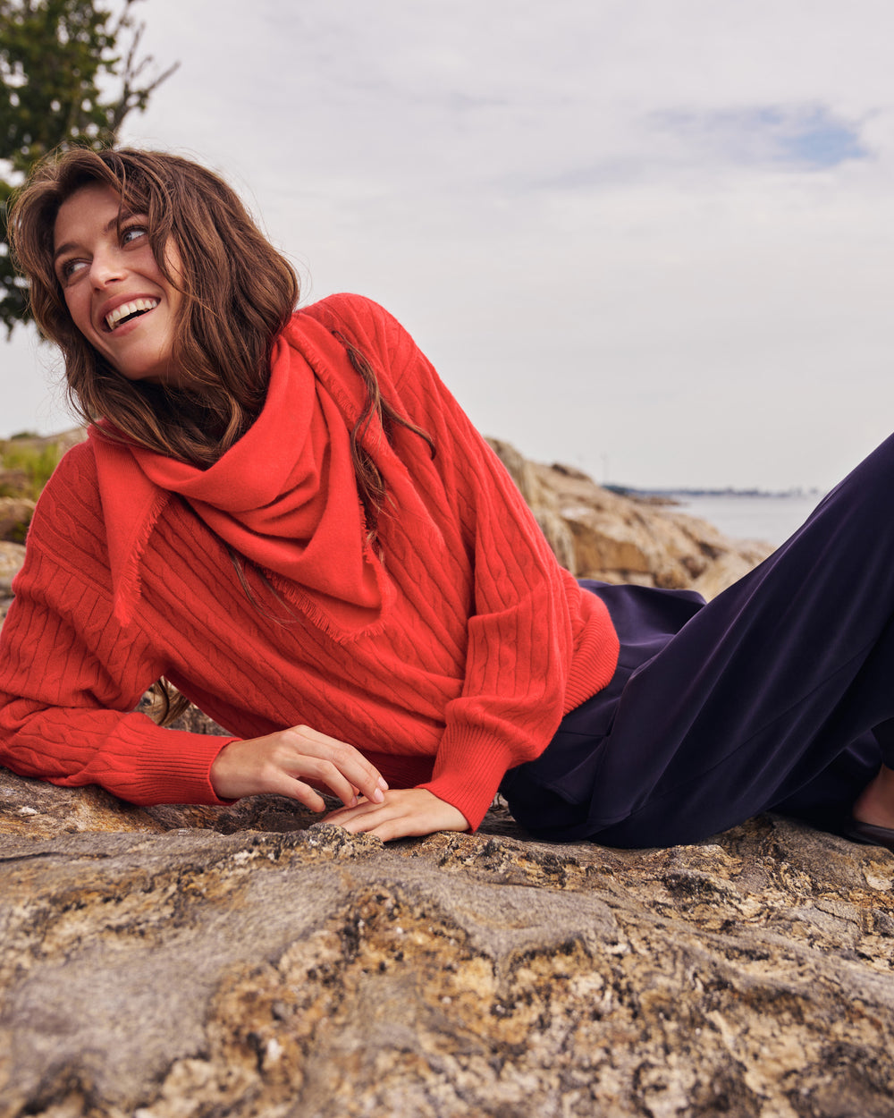 A woman lying on a rocky beach wearing a bright coral-red cable-knit cowl-neck sweater and dark navy pants, with the ocean and cloudy sky in the background.
